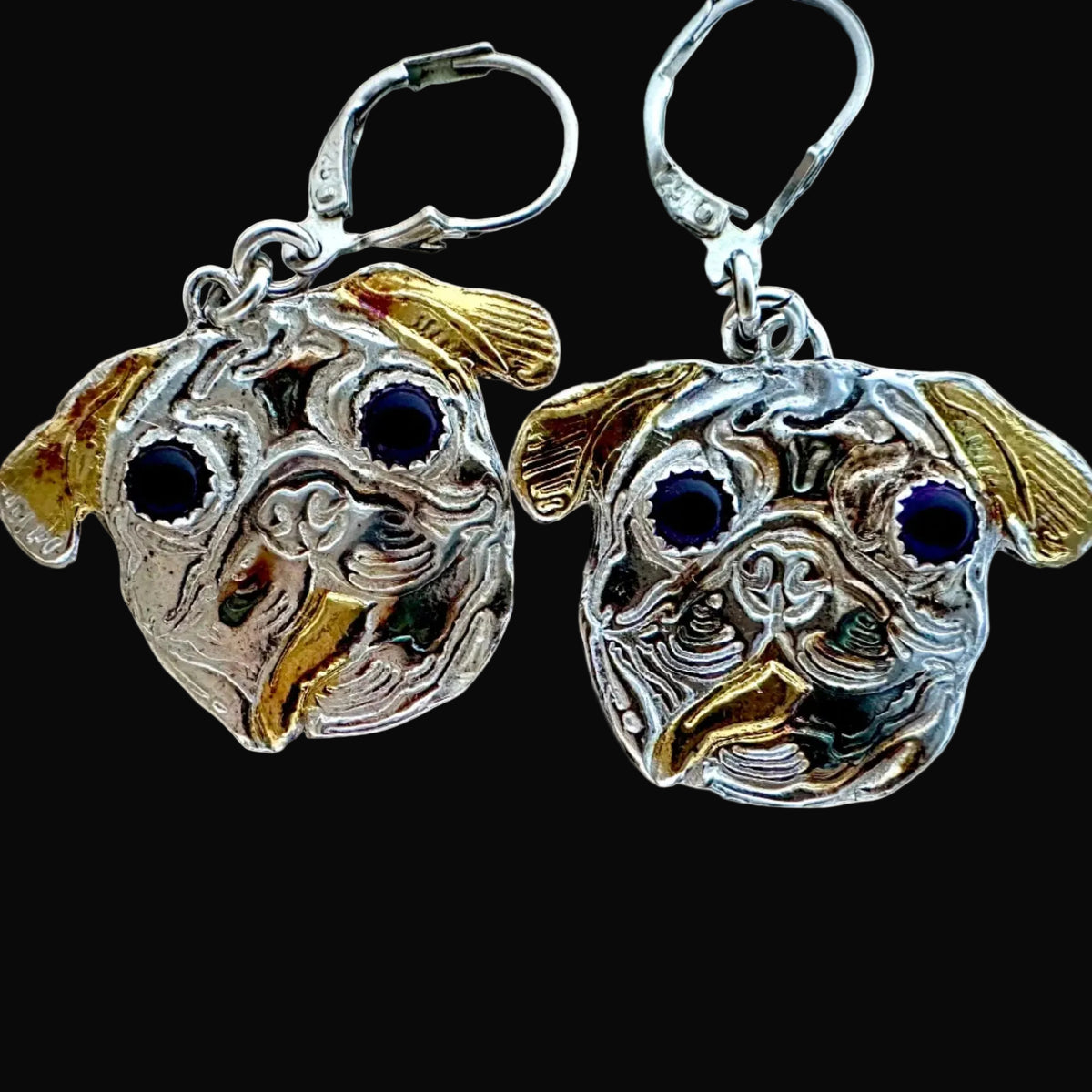 A pair of sterling silver and gold dog face drop earrings with dark blue stone eyes, shown against a solid black background.